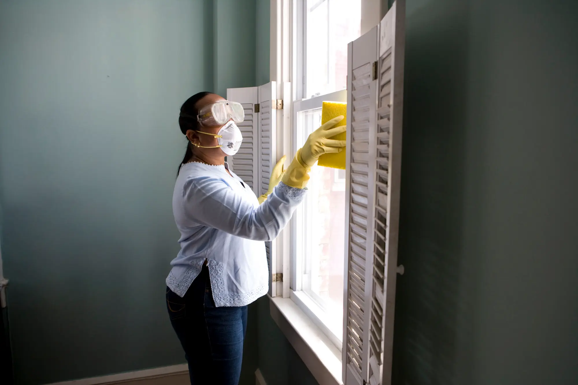 A woman is cleaning the window while wearing a pair of waterproof gloves, a face mask, and a pair of goggles. She is working on her house cleaning in philadelphia pa and is doing a very thorough job